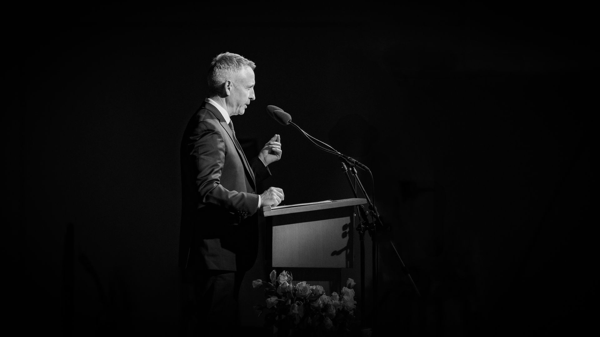 Man in suit speaking at a podium with microphones, under a spotlight. Black and white photograph.