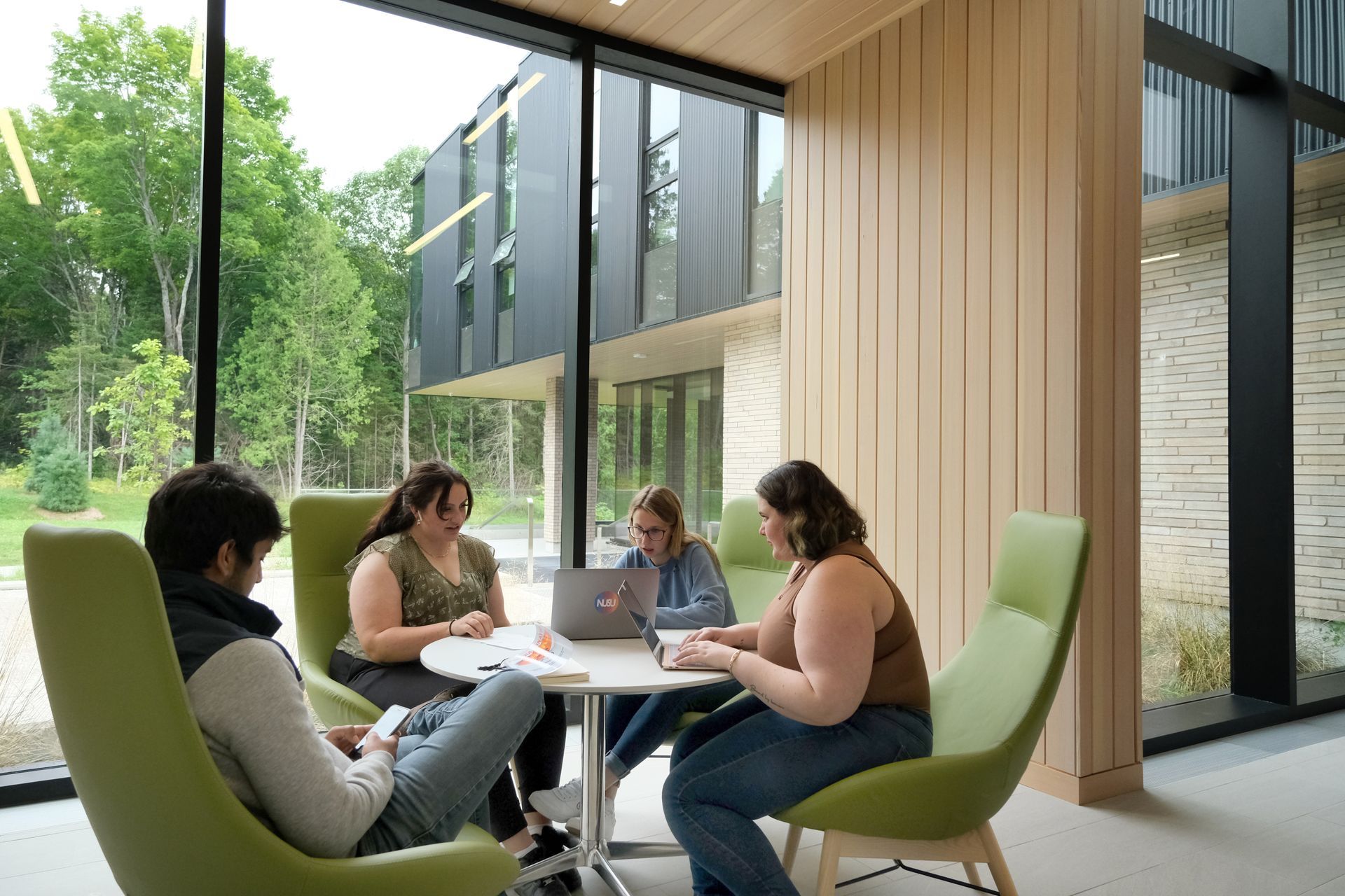 Four people seated around a table, in a room with large windows, green chairs, and a view of trees.