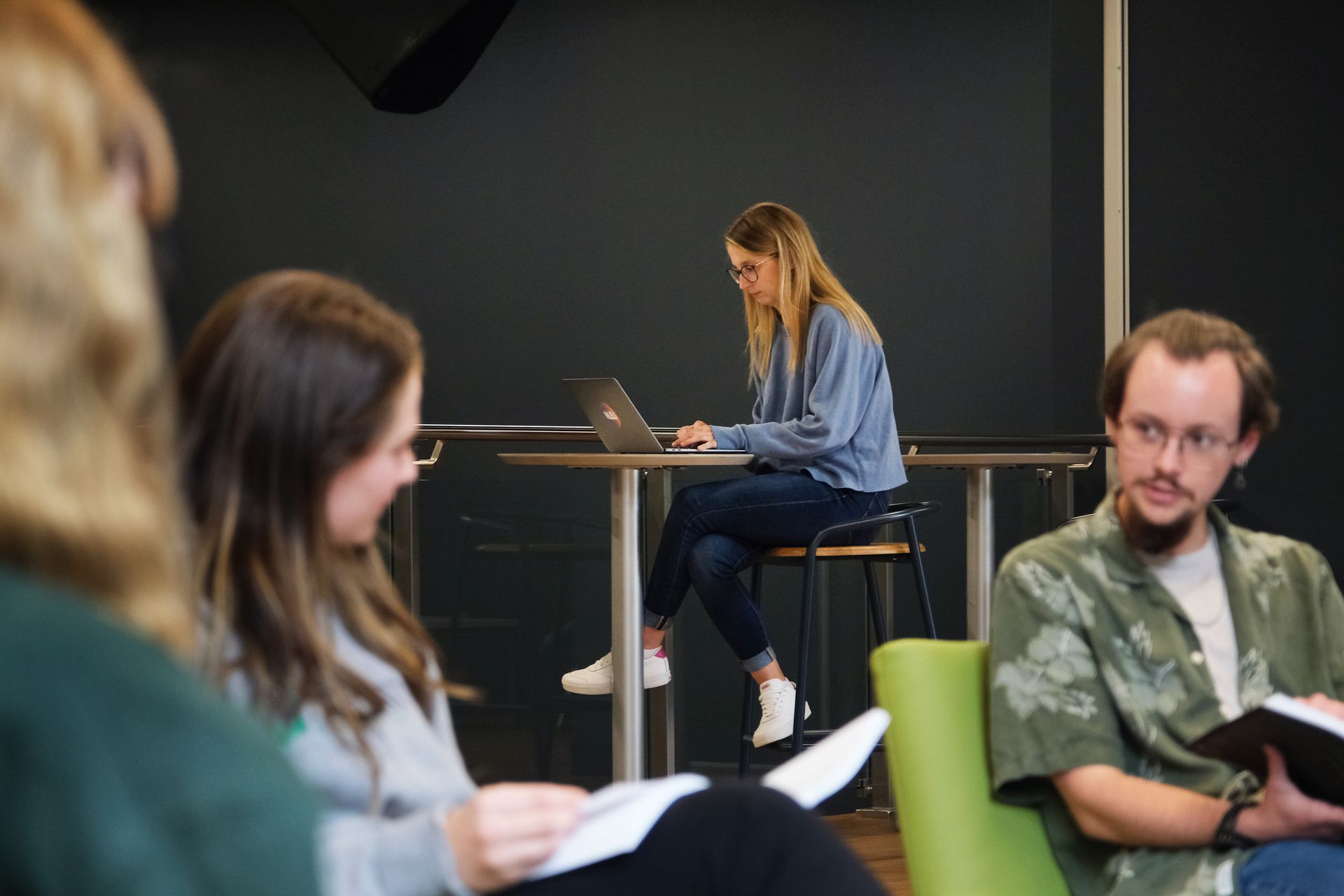Four students in a room. One woman works on a laptop, while others read. Dark backdrop, modern setting.