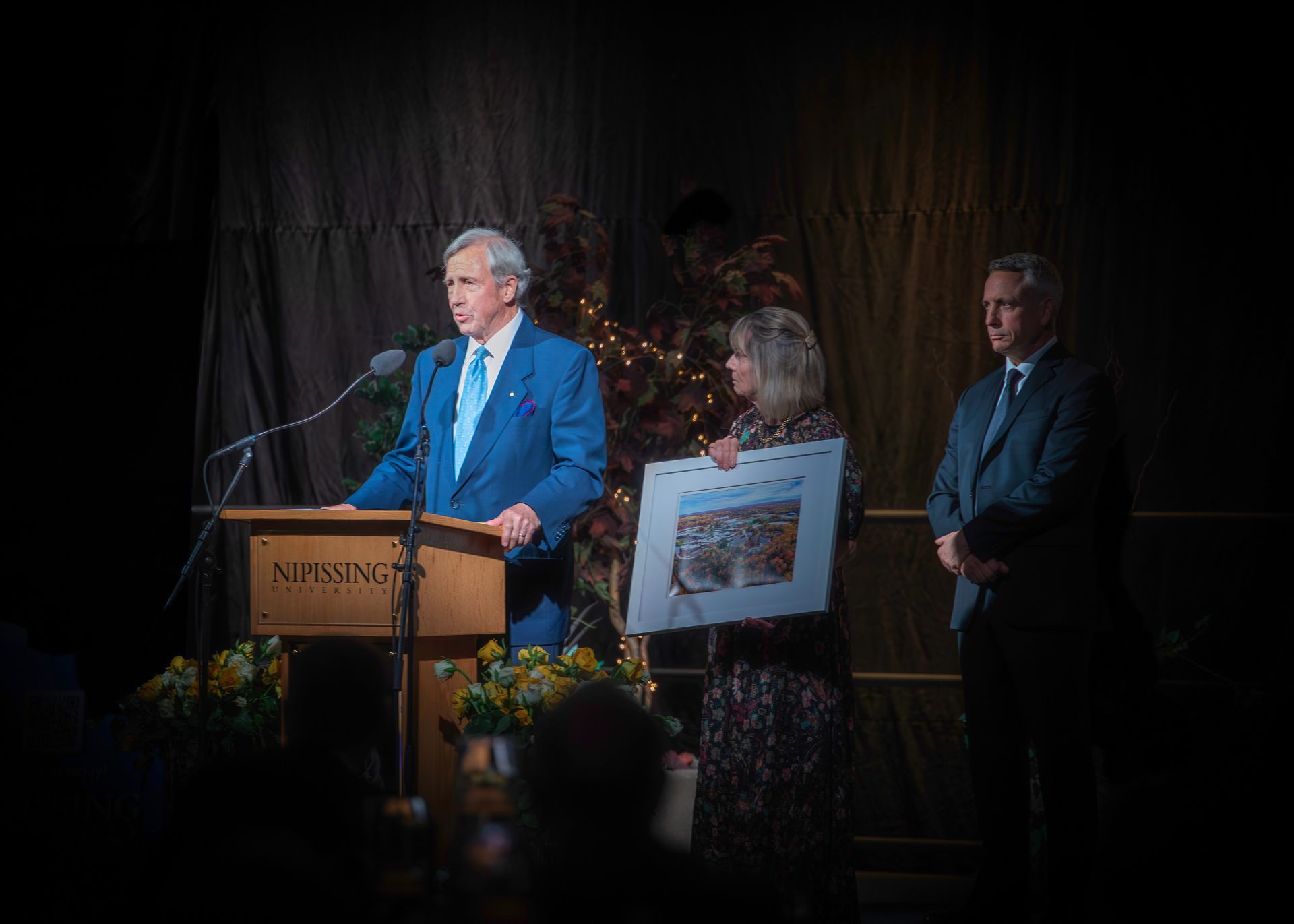 Man in blue suit speaking at a podium, two people stand beside him holding a framed picture in a dimly lit setting.