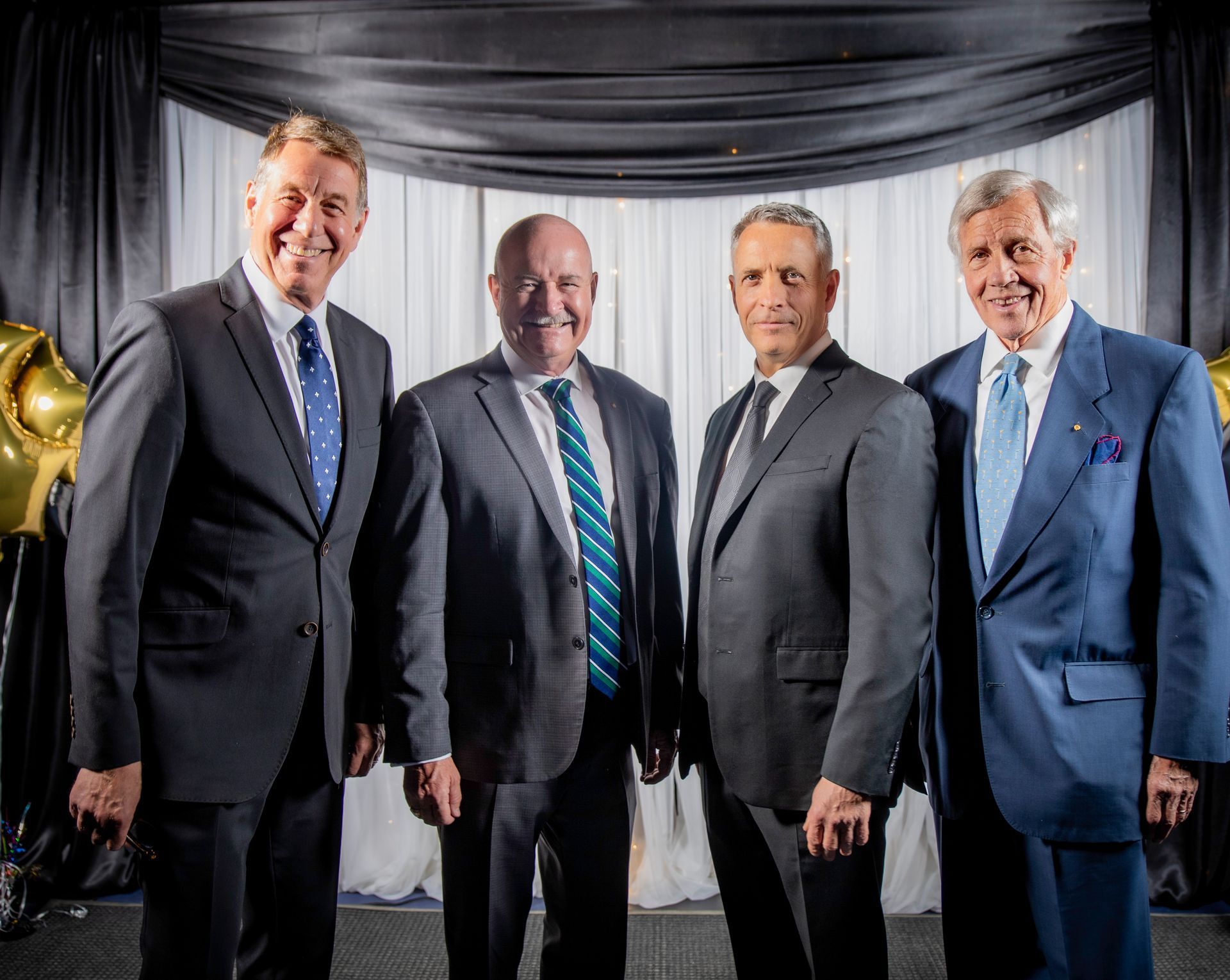 Four men in suits stand together in front of a draped backdrop. They smile for a group photo.