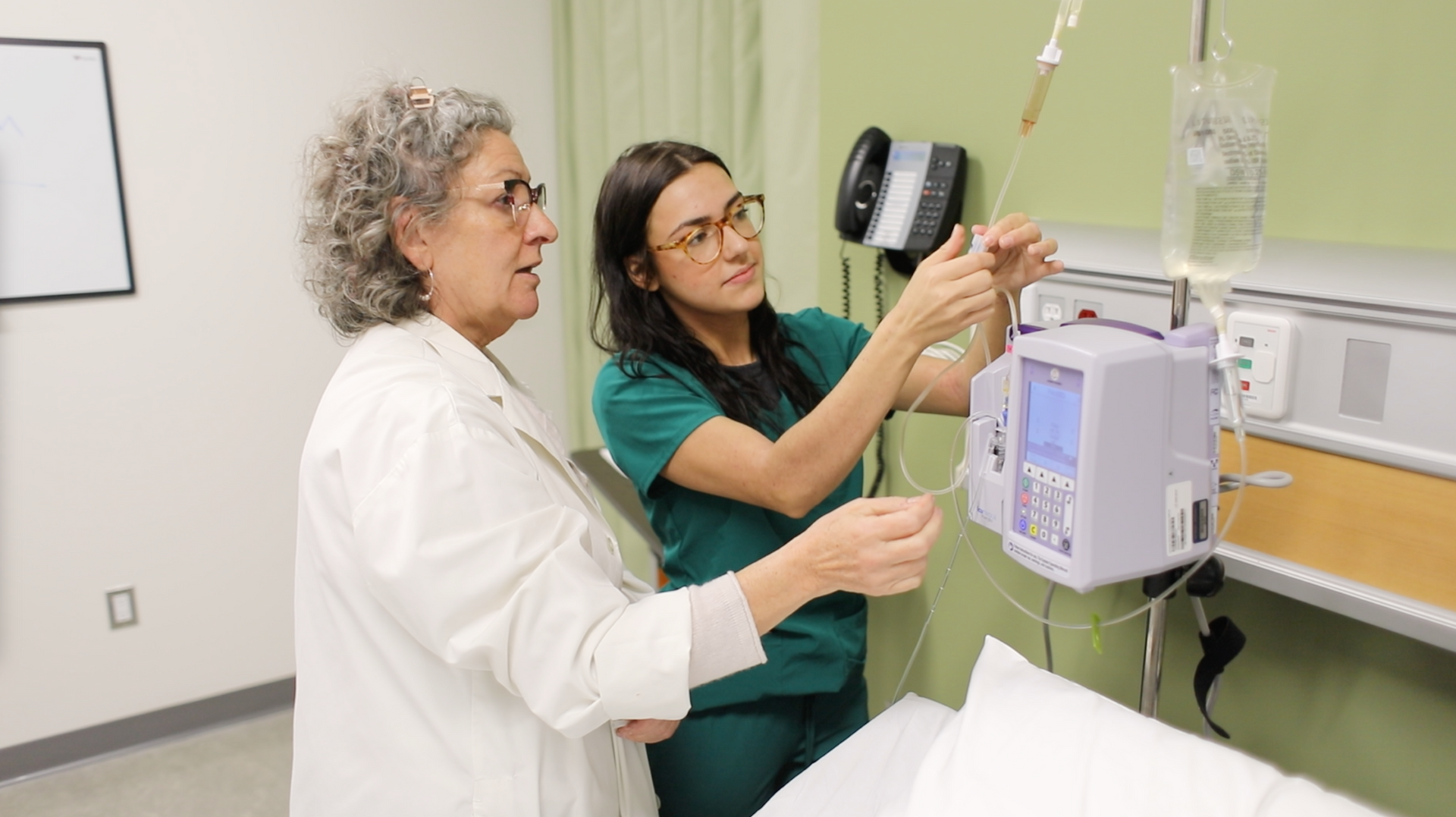 Two healthcare workers adjusting an IV bag and machine in a hospital room.