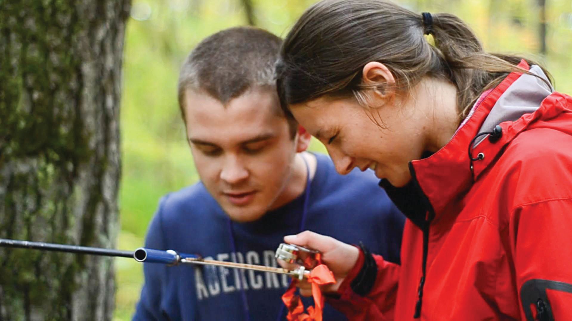 Man and woman outdoors, looking at gear, red jacket, blue shirt, forest background.