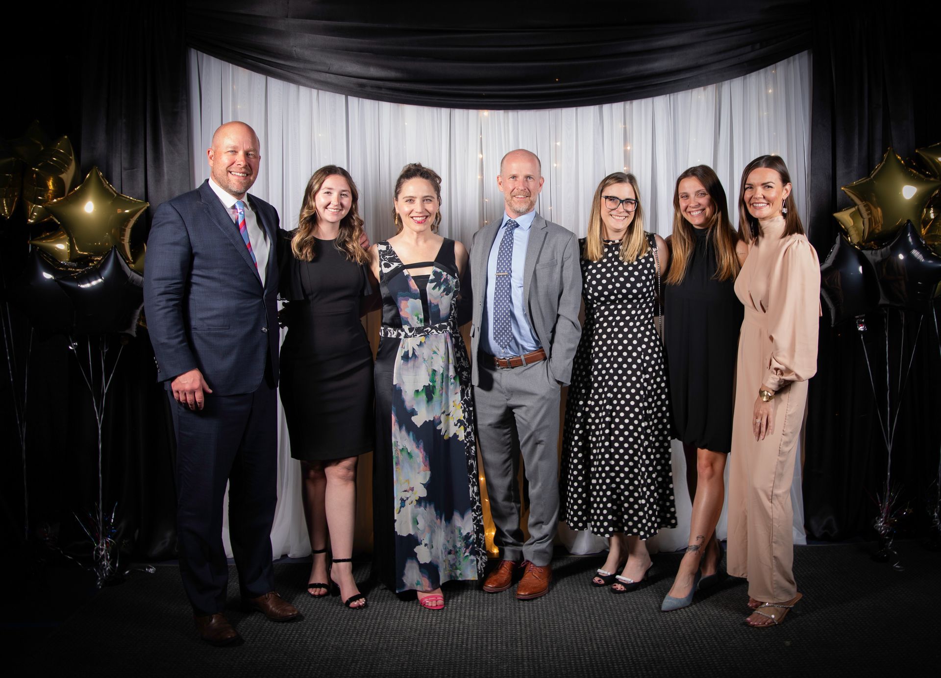 Group of seven people smiling at a formal event with a white backdrop.