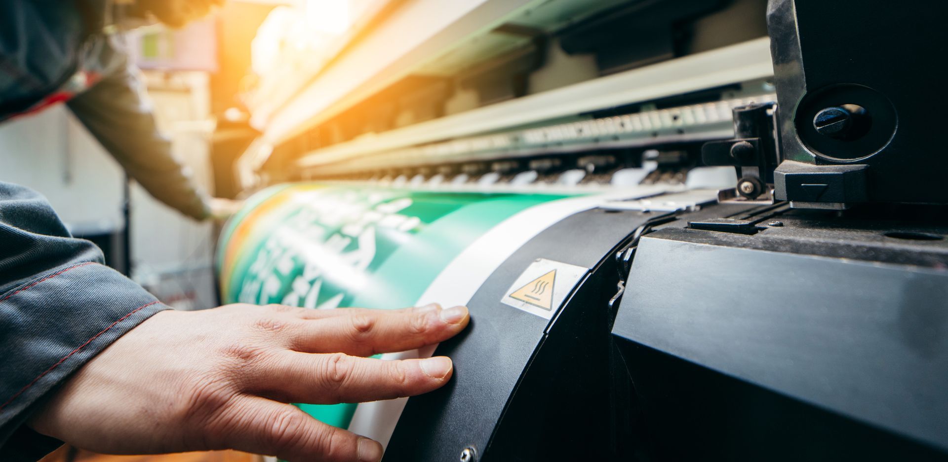 Person's hand on a large format printer printing a green banner with white text.