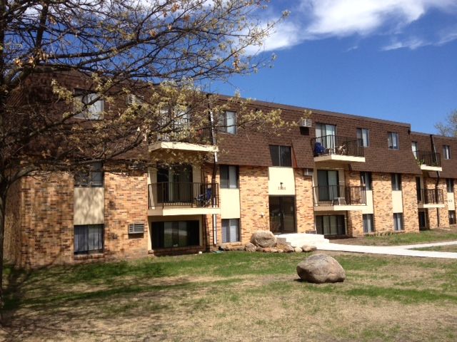 Apartment building with brown roof and brick exterior. Green lawn in front, sunny sky.