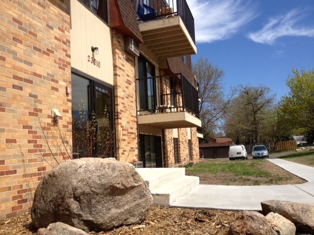 Apartment building with brick facade and balconies, sunny day. Large rocks in foreground.