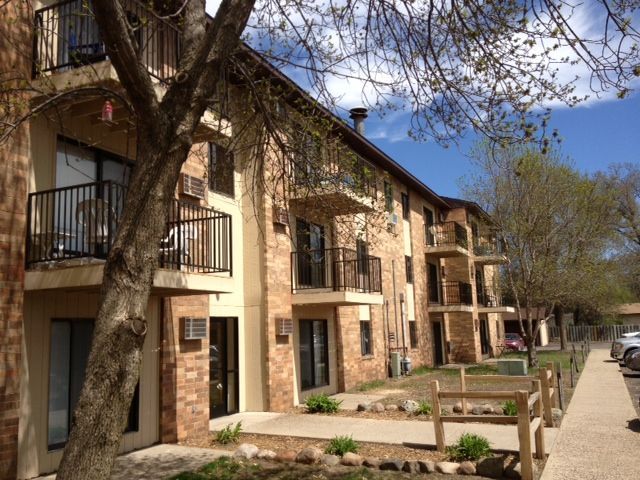 Apartment building with brick facade, balconies, and a tree in front on a sunny day.