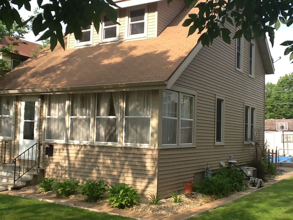 Two-story beige house with a brick facade, porch, and brown roof. Lush green plants line the foundation.