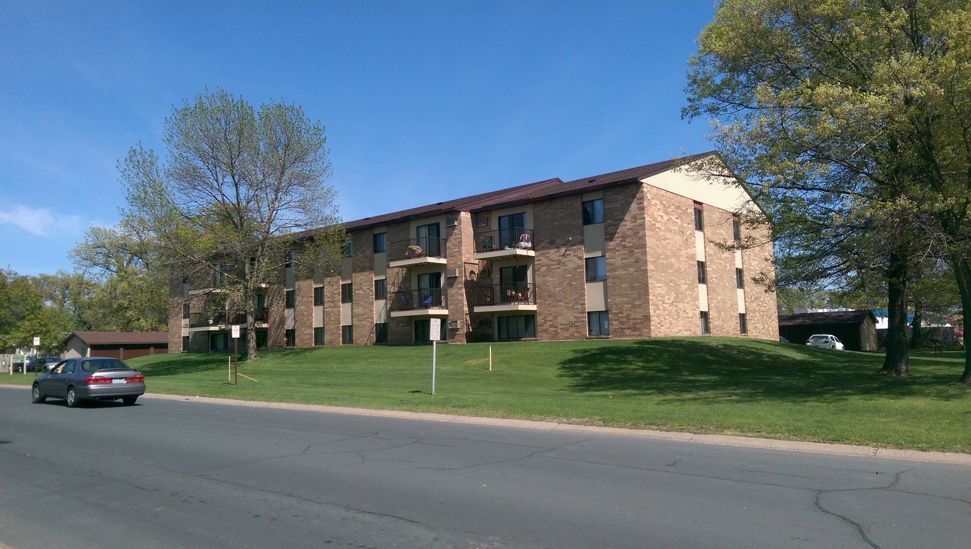 Apartment building with balconies on a grassy hill next to a road; car driving by.
