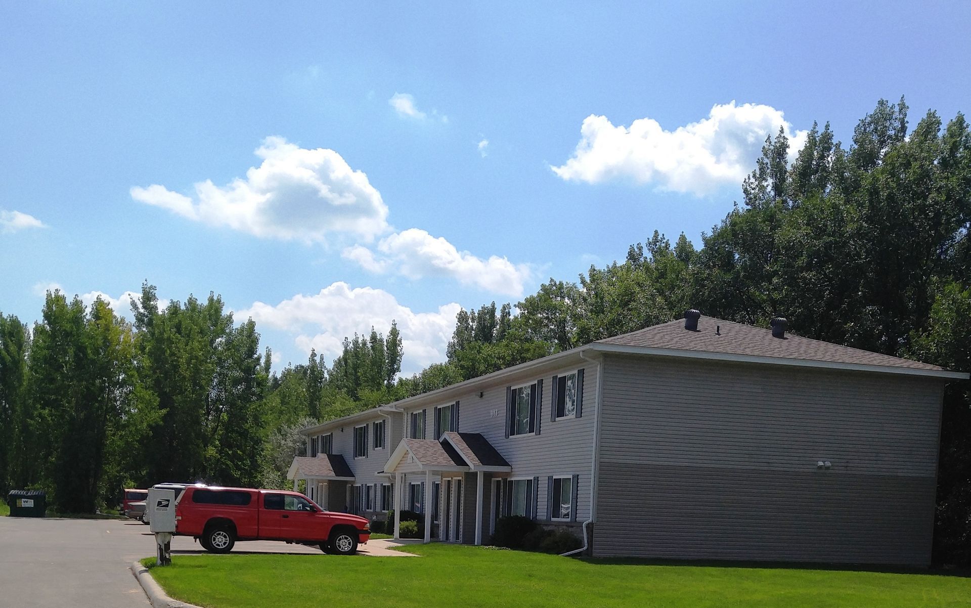 Two-story apartment building with a red pickup truck parked in front. Green grass and trees under a blue sky with clouds.