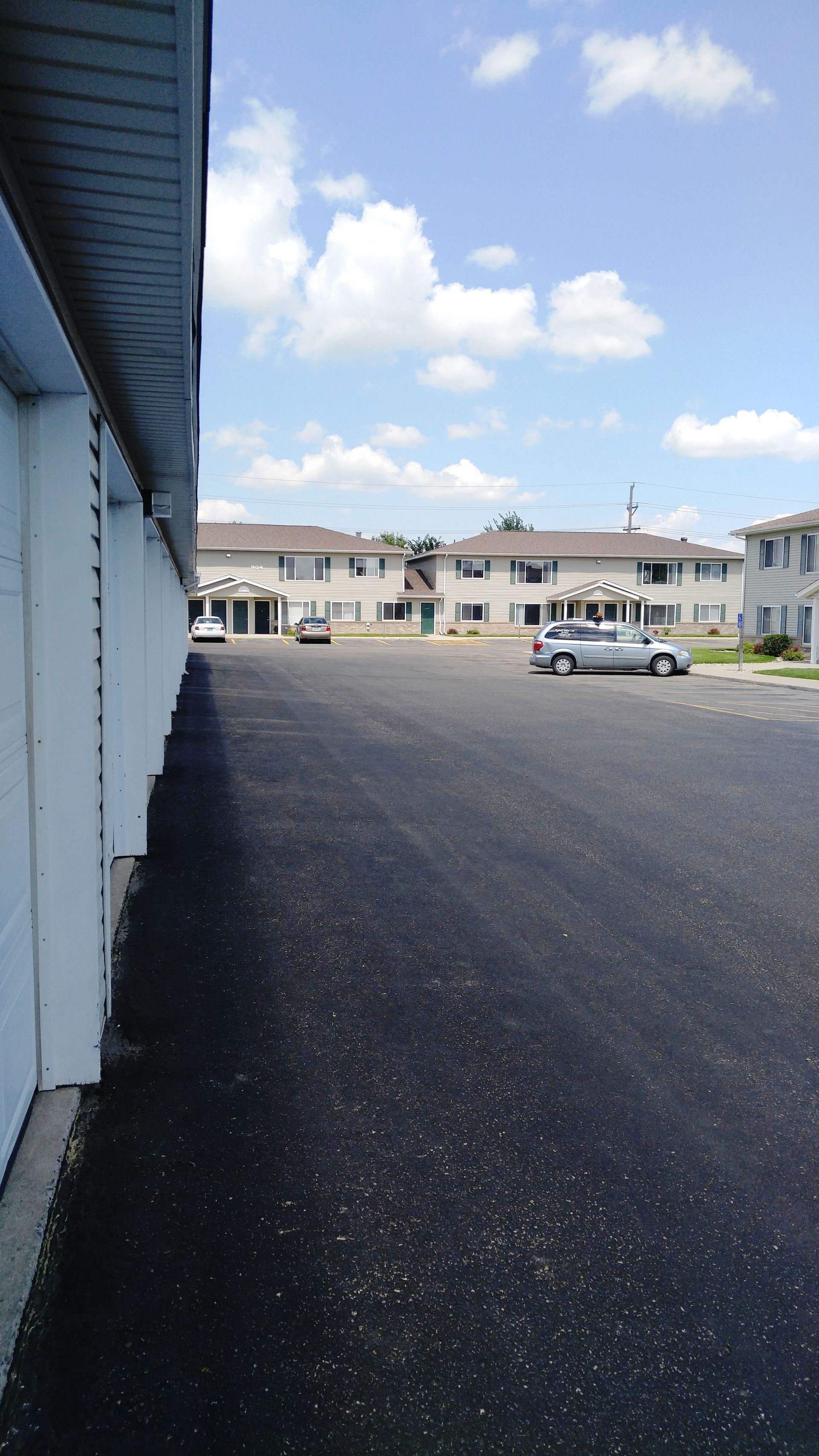 Apartment complex with long asphalt parking lot under a blue sky with white clouds.