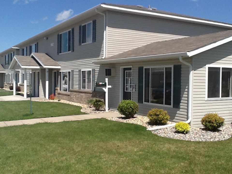 Two-story townhouses with gray siding, green shutters, and manicured lawns under a blue sky.