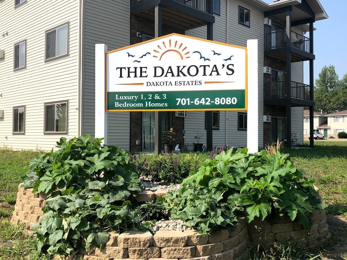 Sign for The Dakota's Dakota Living apartments, with a phone number and unit info, in front of a building and landscaping.