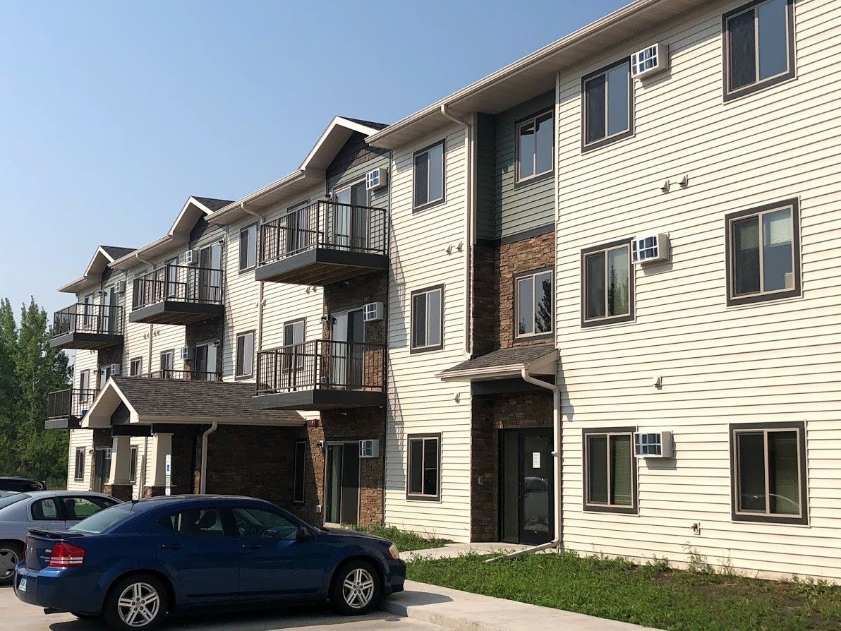 Apartment building exterior with balconies and parked cars. Blue sky. Beige siding with brick accents.