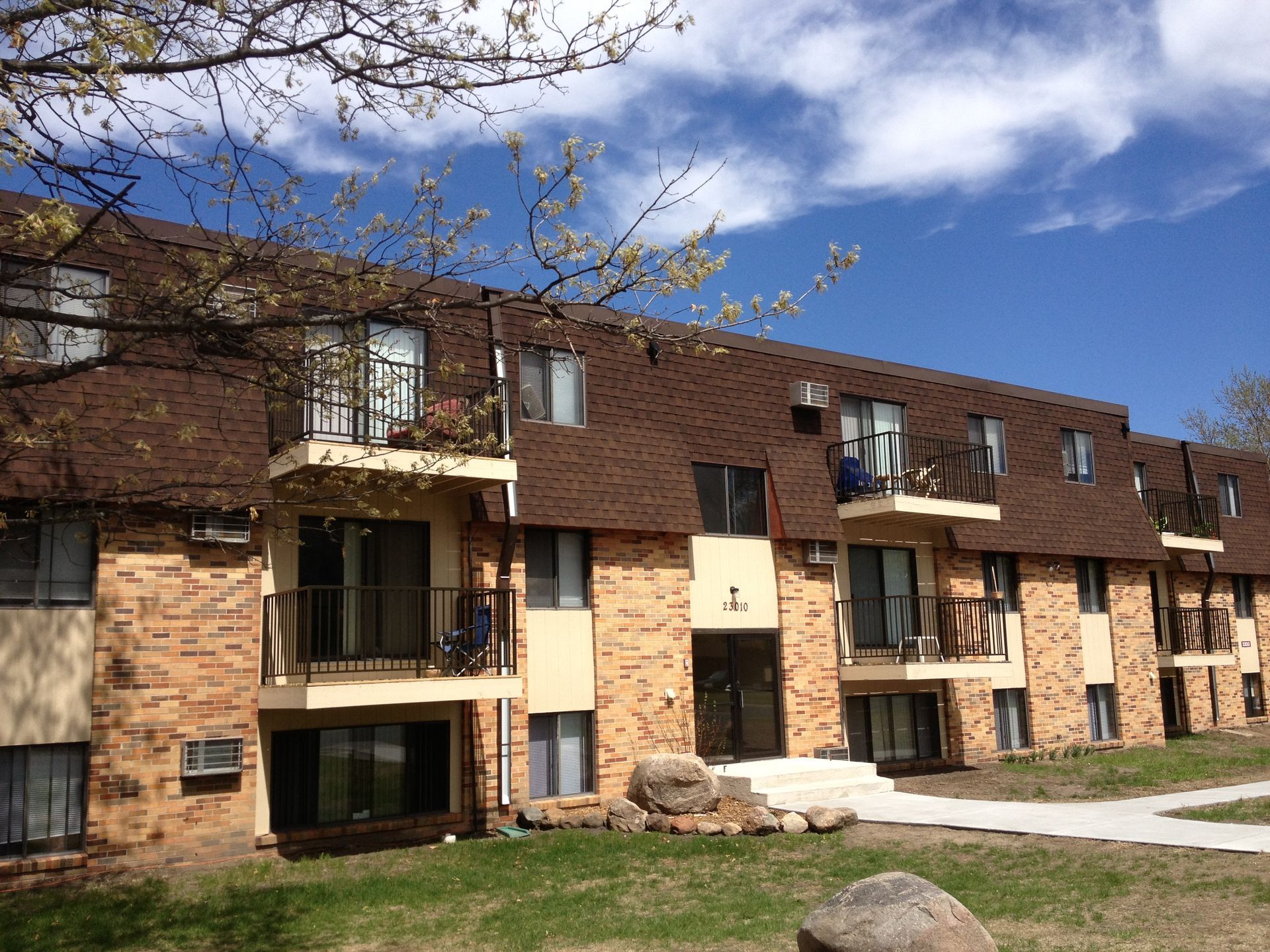 Two-story apartment building with brown roof, tan brick and siding, balconies, and green grass on a sunny day.