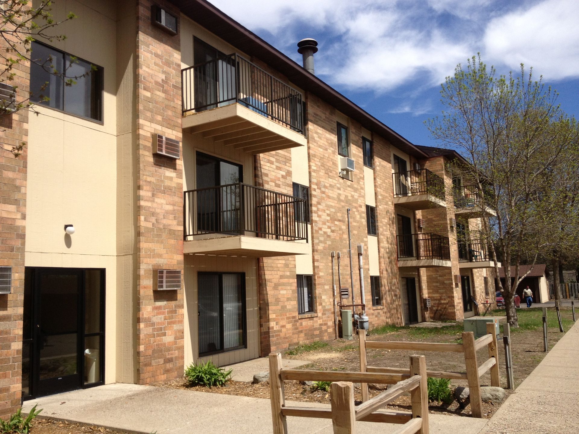 Multi-story brick apartment building with balconies, sidewalk, and small wooden fence.