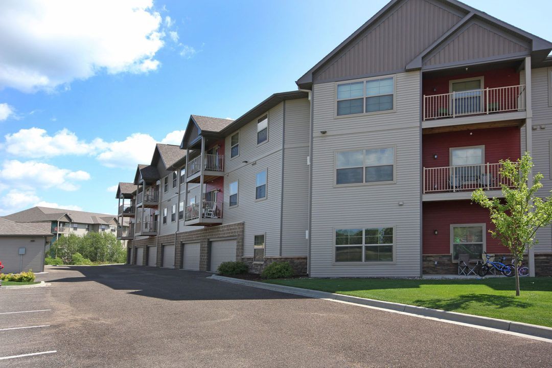 Apartment building with balconies, red accents, garages, and parking area, on a sunny day.