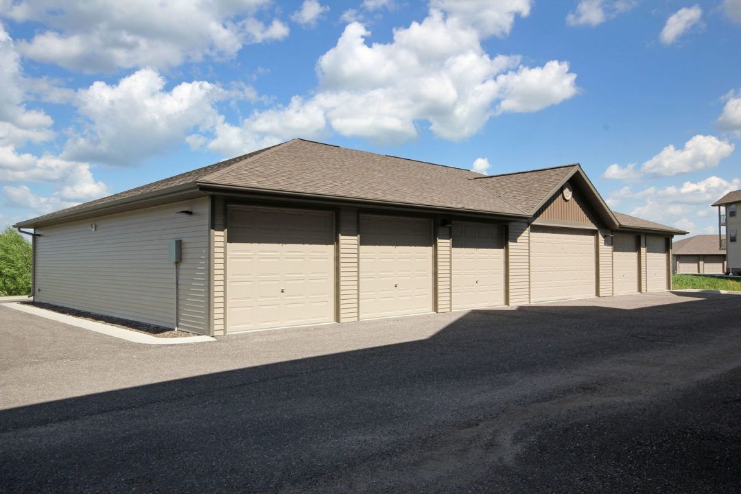 Row of tan garage doors on a beige building under a brown roof with blue sky and clouds overhead.