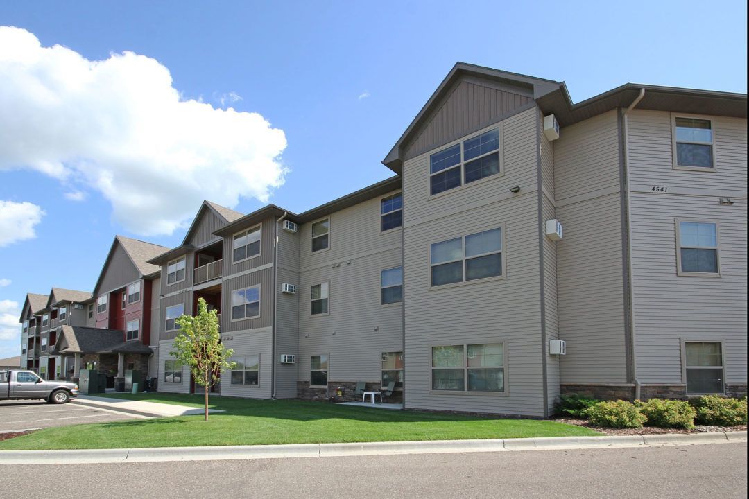 Apartment building with gray siding, brown accents, blue sky, and patches of green grass.