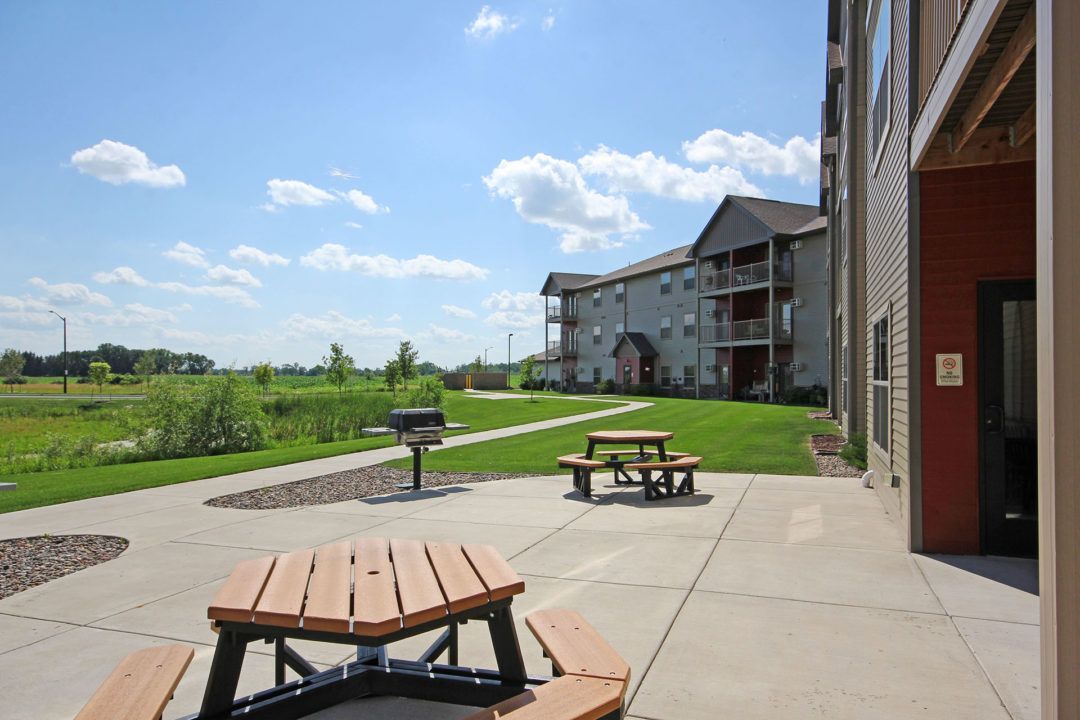 Outdoor patio with picnic tables, grill, and apartment building under a blue sky.