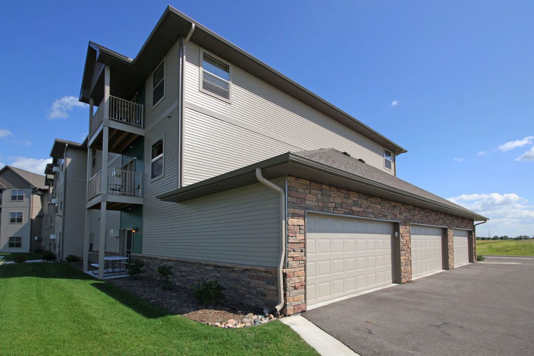 Apartment building with tan siding and stone accents, garages, green lawn, and blue sky.