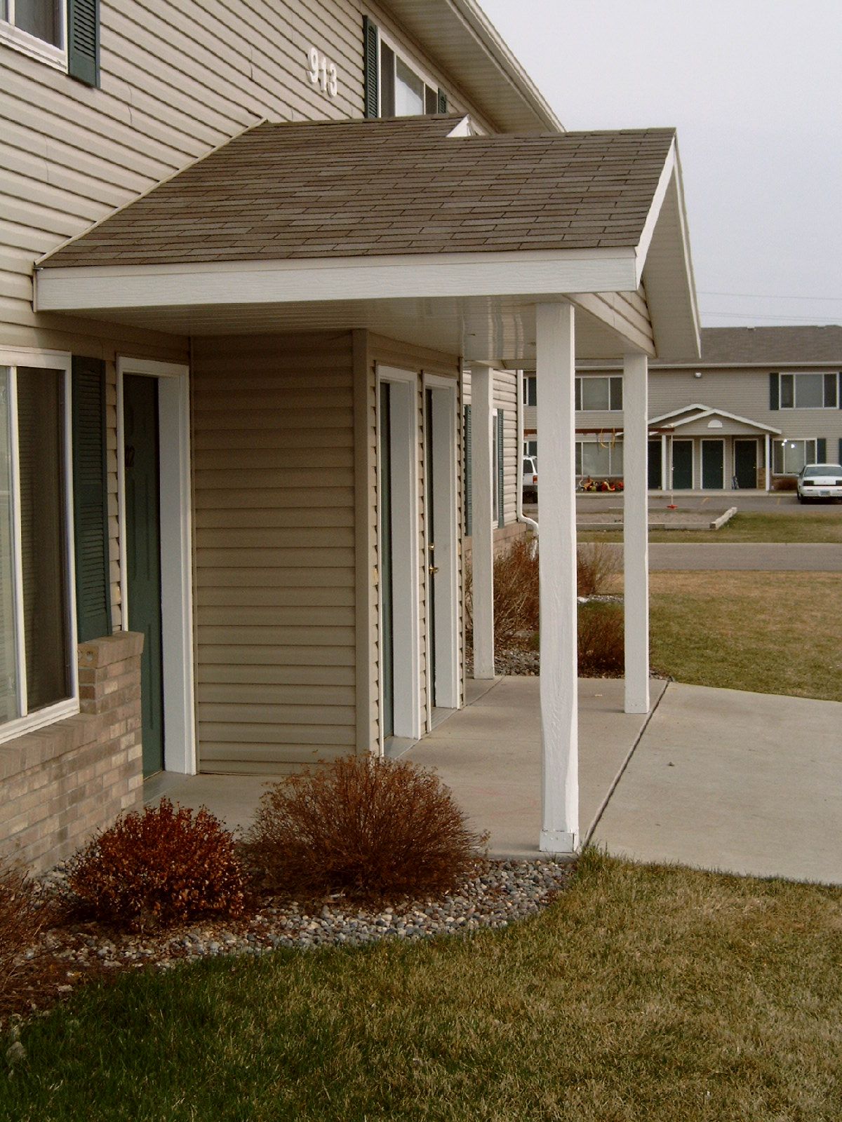 Apartment building with covered walkway, white columns, tan siding, brown roof, concrete path, and green grass.