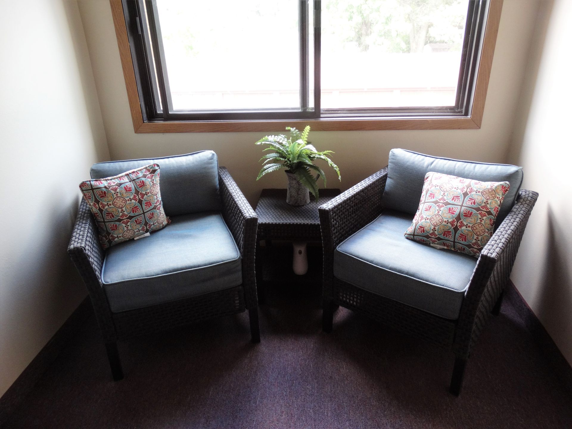Two blue wicker chairs with pillows and a small table with a plant by a window.