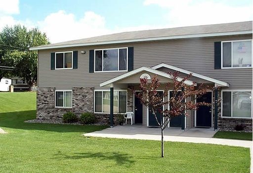 Two-story apartment building with tan siding, stone accents, and green lawn.