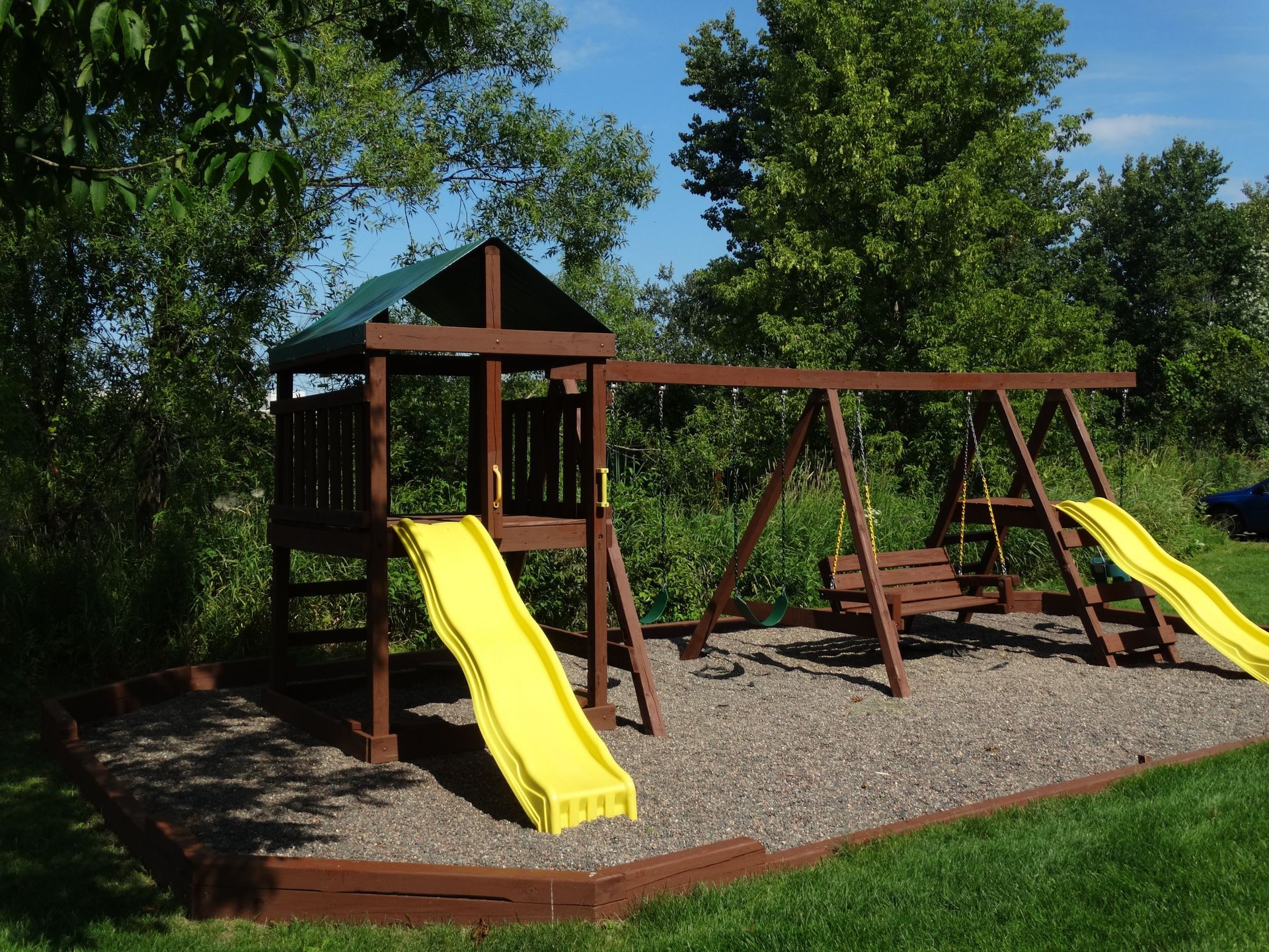 Wooden playground with a slide, swings, and a covered platform on gravel, in a yard.