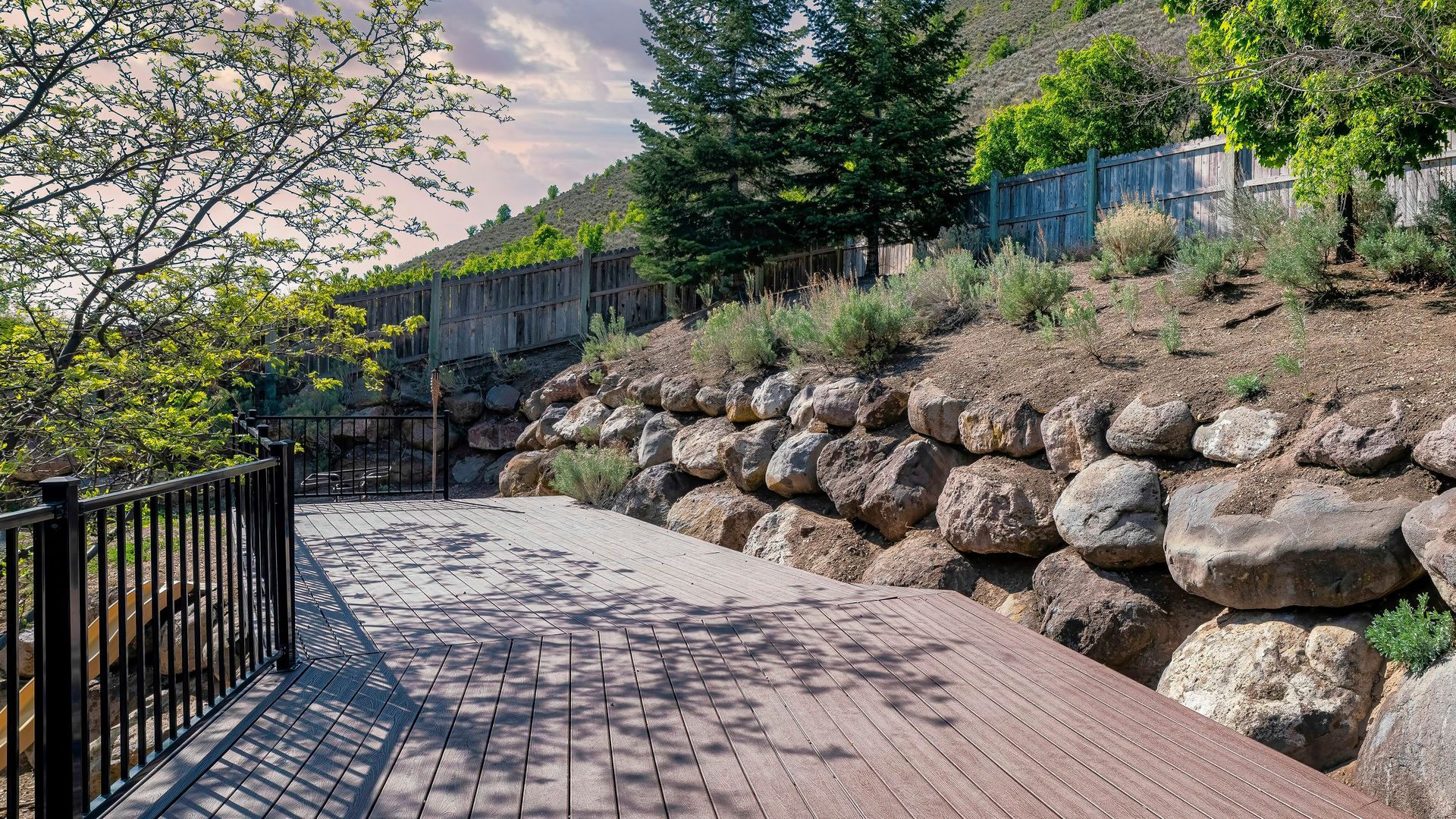 Wooden walkway with black railing, rock retaining wall, and fenced hillside.
