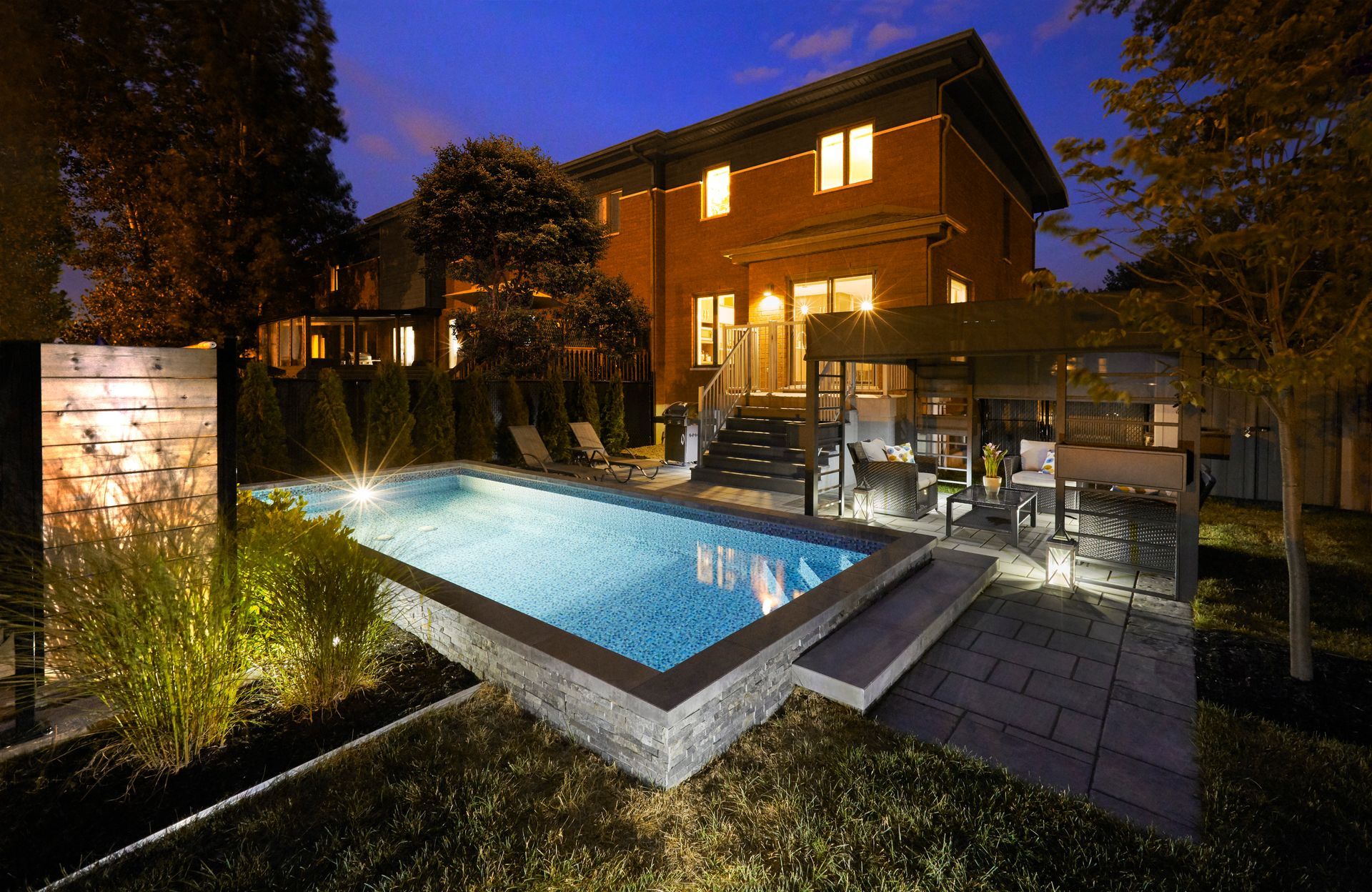 Backyard at night with illuminated raised beam pool, patio, and two-story brick house.