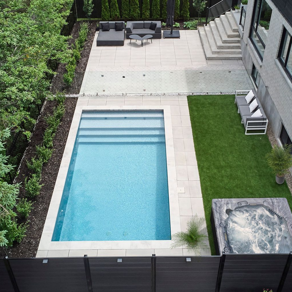 Backyard with pool, patio, and modern house. Lush greenery surrounds the space; the pool has blue tile.