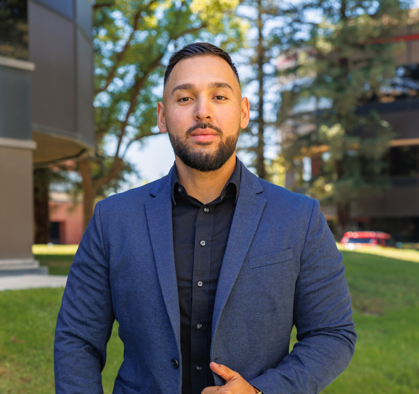 A man in a blue jacket and black shirt is standing in front of a building.