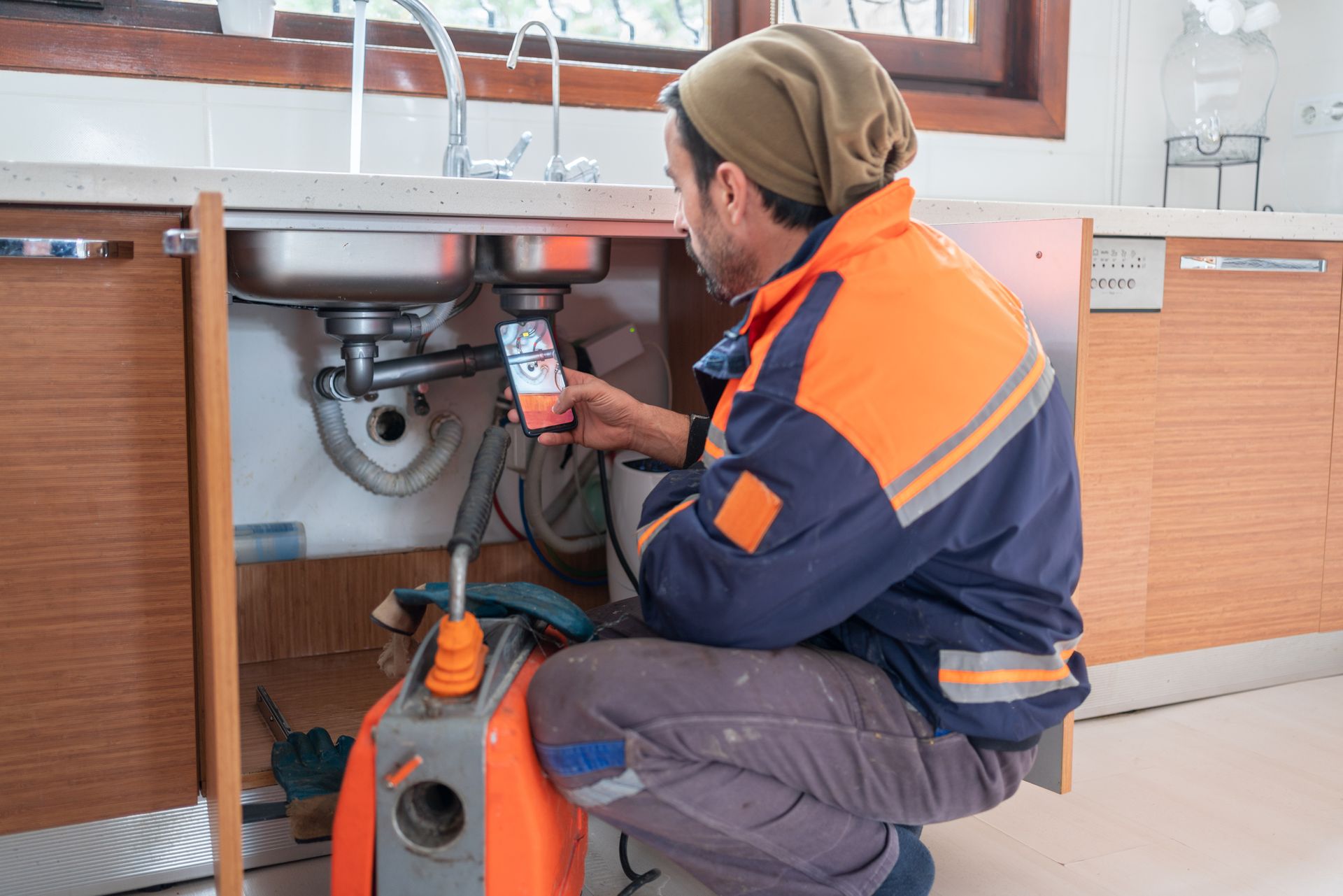 A plumber is kneeling down under a sink in a kitchen.