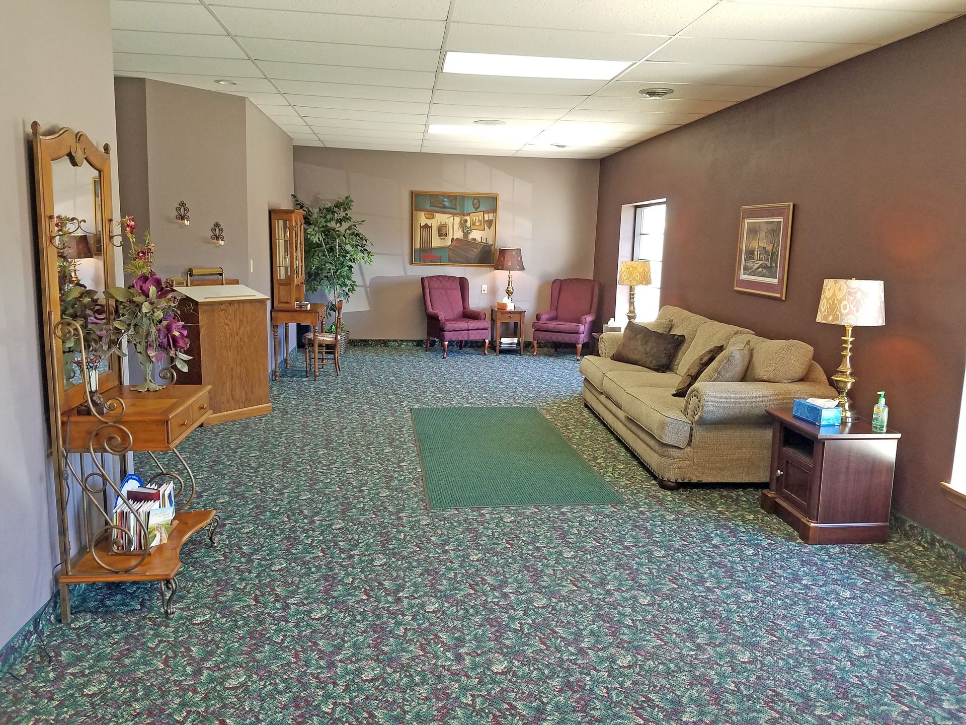 Lobby with floral carpet, mauve and dark red walls, two purple armchairs, a couch, and a tall plant.