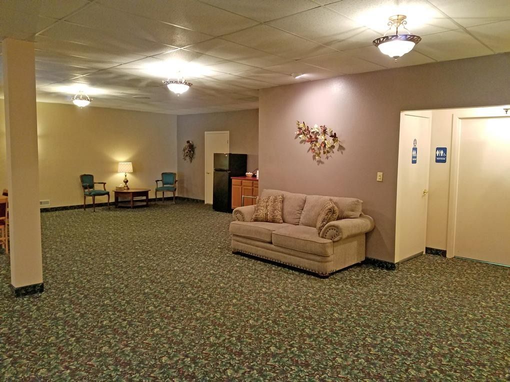 Spacious room with floral carpet, a sofa, chairs, and a small fridge. Neutral walls and overhead lighting.