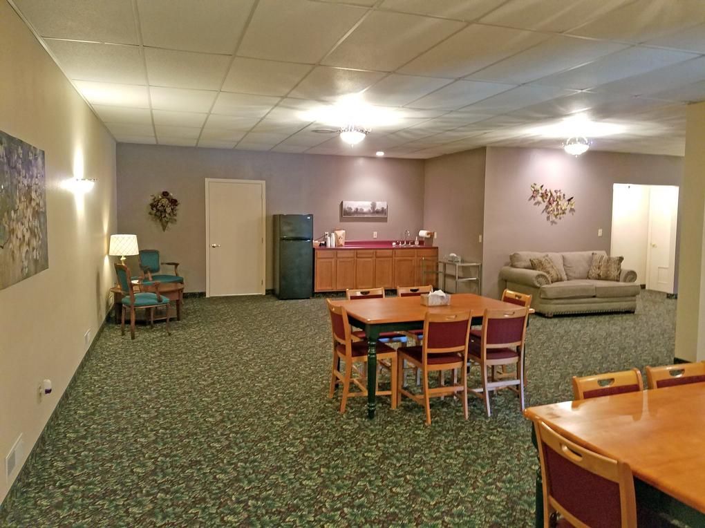 Large room with tables, chairs, sofa, cabinets, refrigerator, and decorative wall art. Green carpet, neutral walls.