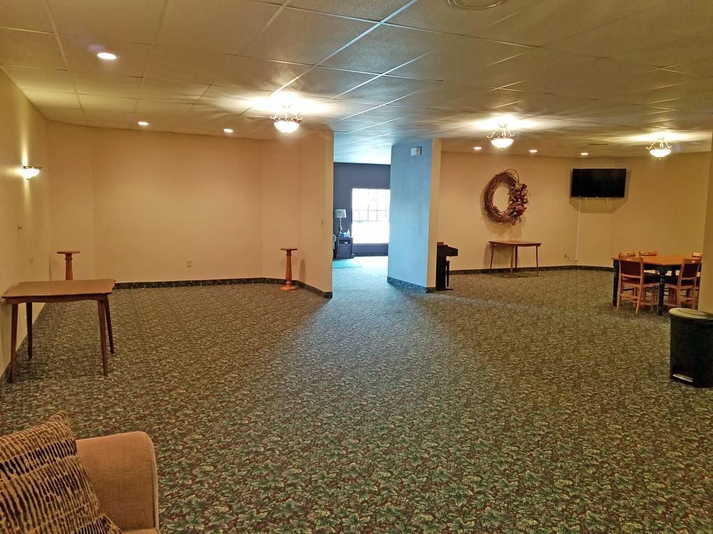 Spacious room with patterned carpet, tan walls, tables, wreath, TV, and open doorway to exterior.