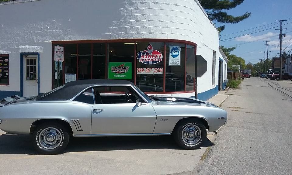 A silver classic Chevrolet Camaro parked on the street in front of a white brick building on a sunny day.