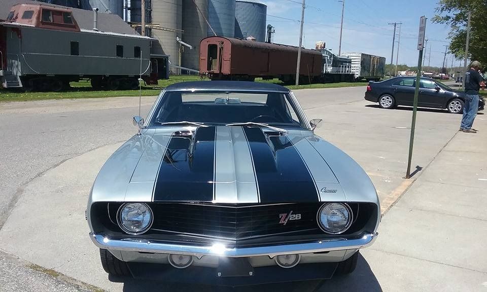 A silver Chevrolet Camaro with black racing stripes parked on a street near train cars on a sunny day.