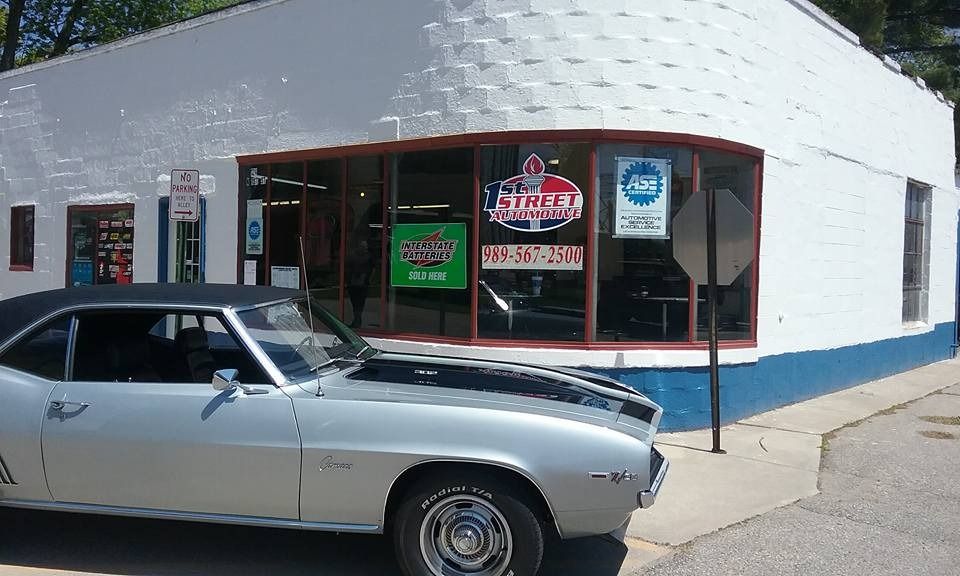 A silver 1968 Camaro parked in front of a white and blue corner building with storefront windows displaying signs.