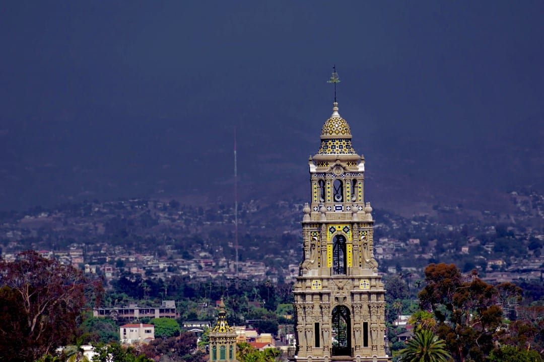 A large clock tower with a cross on top of it is in the middle of a city.
