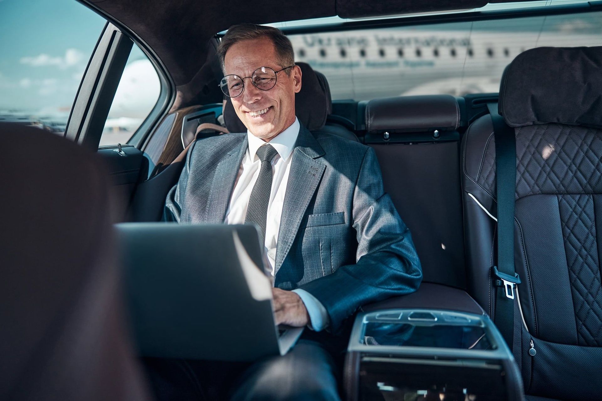 A man in a suit is sitting in the back seat of a car using a laptop computer.