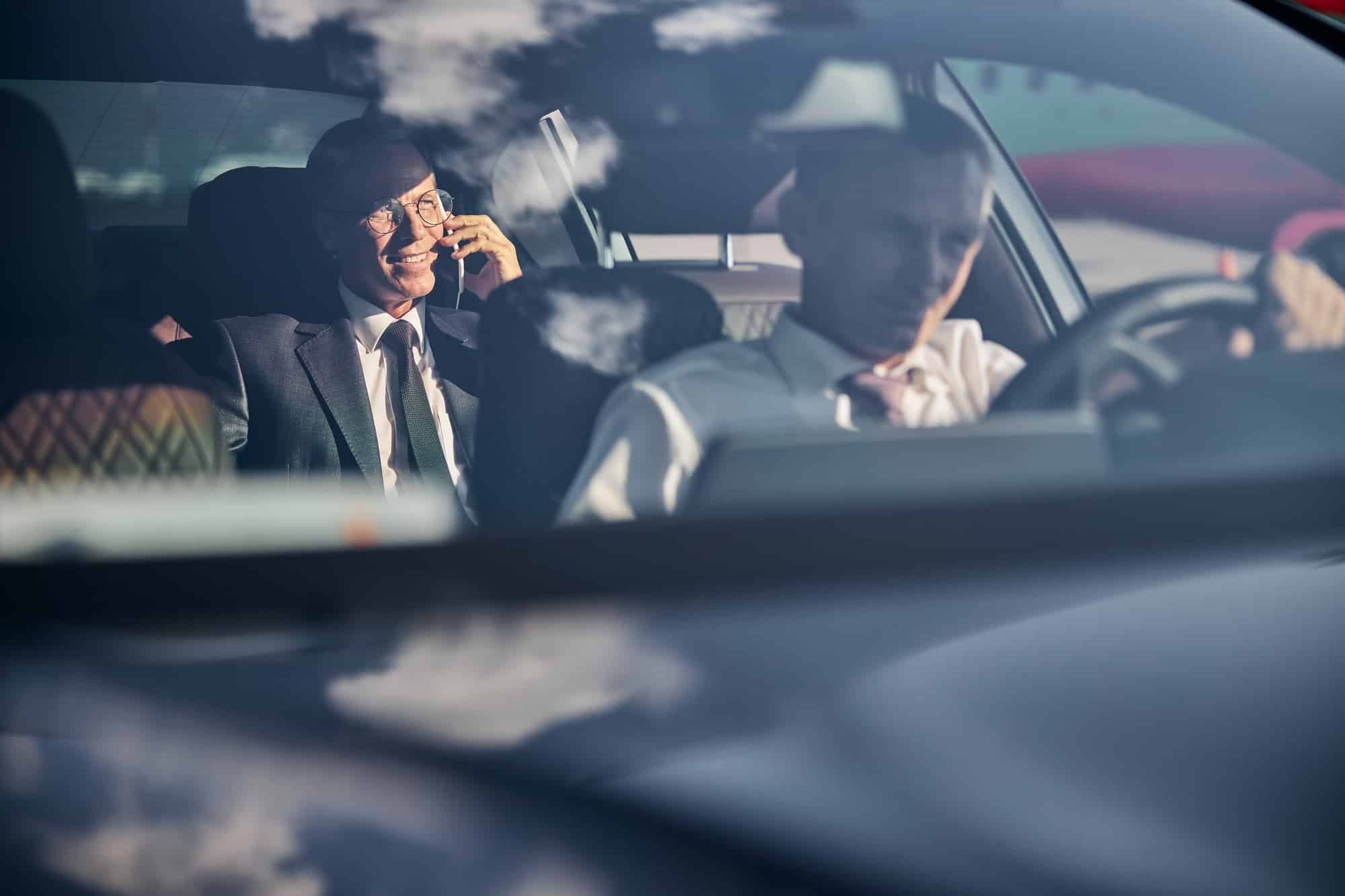 Two men are sitting in a car talking on cell phones.