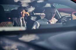 Two men are sitting in a car talking on cell phones.