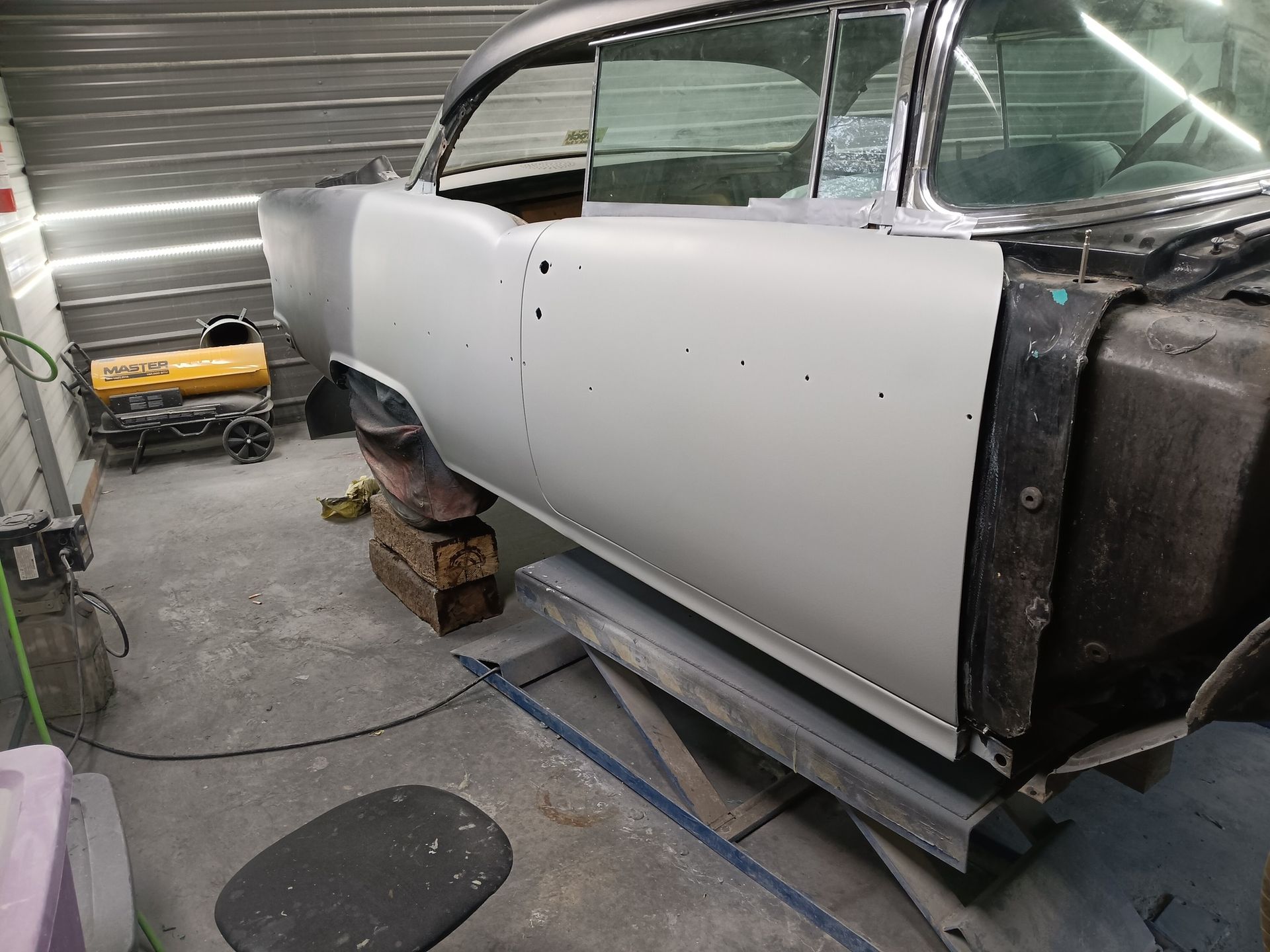 A partially restored classic car body painted in gray primer sits on a metal lift in a workshop.