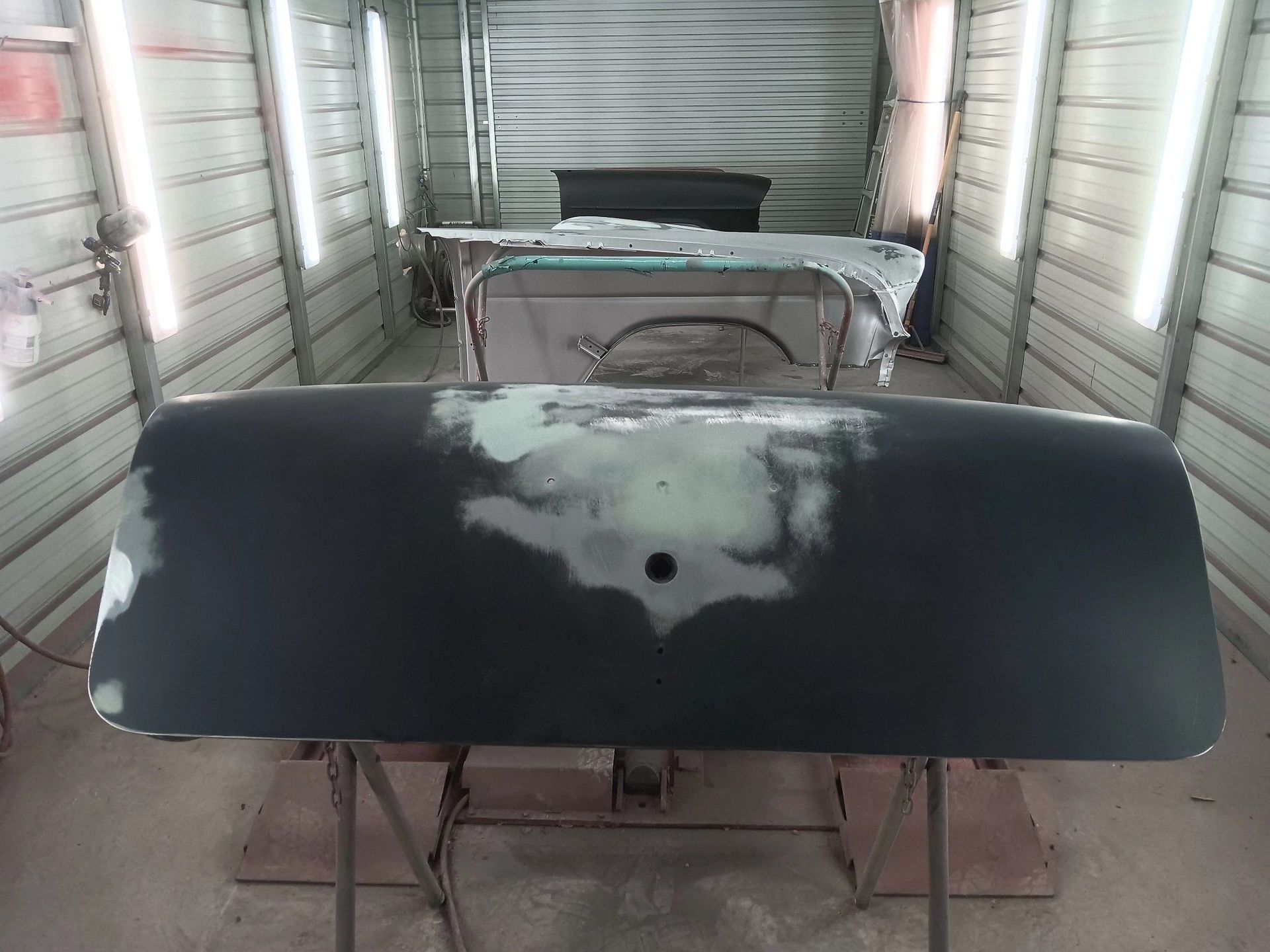 A matte black car door mounted on a stand inside a paint booth, showing sanded body filler patches on its surface.