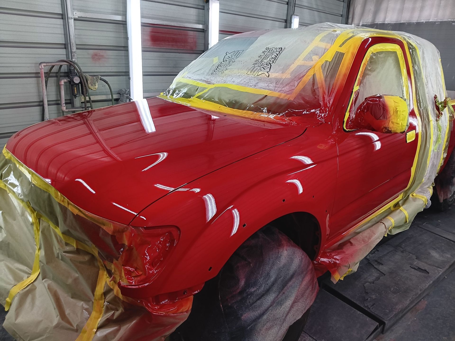 A vibrant red truck sits in a paint booth, with windows and tires masked off with tape and plastic film.