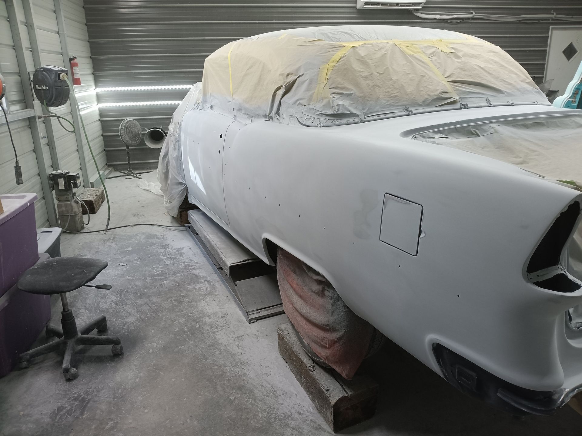 A primer-coated vintage car sits on blocks inside a workshop, with the windows masked off for painting.